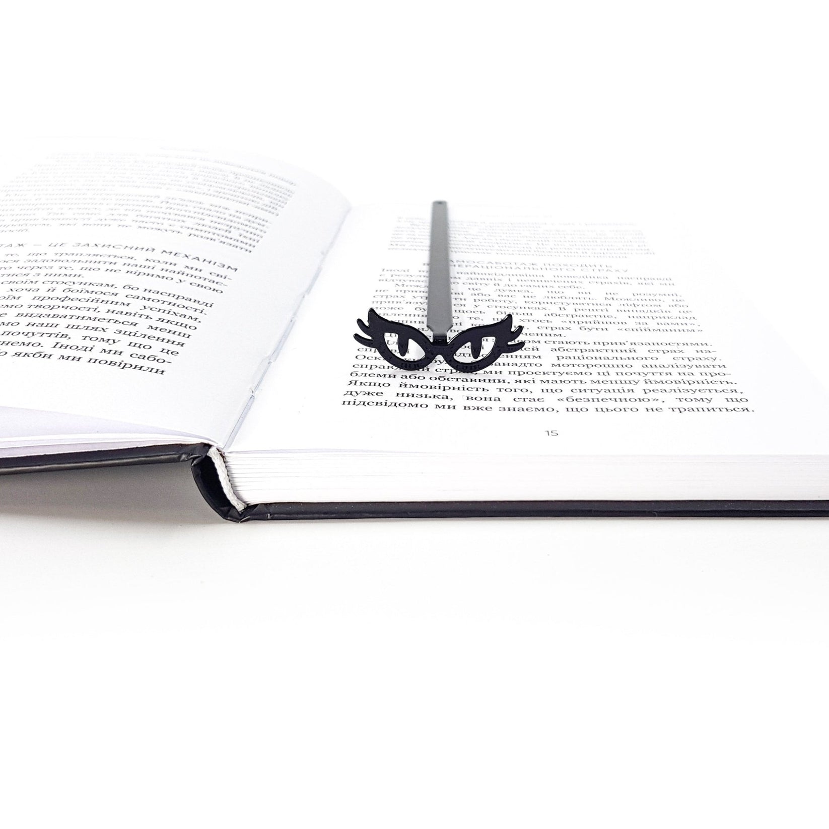 Black metal bookmark shaped like cat’s eye glasses resting in an open book, minimal masquerade-style design marking the page.