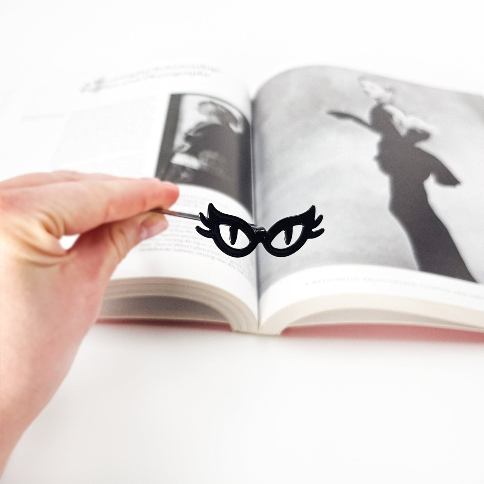 A hand holds a slim black metal bookmark shaped like cat’s eye glasses above an open book, with softly blurred pages in the background, highlighting the playful masquerade-style design against a clean white setting.