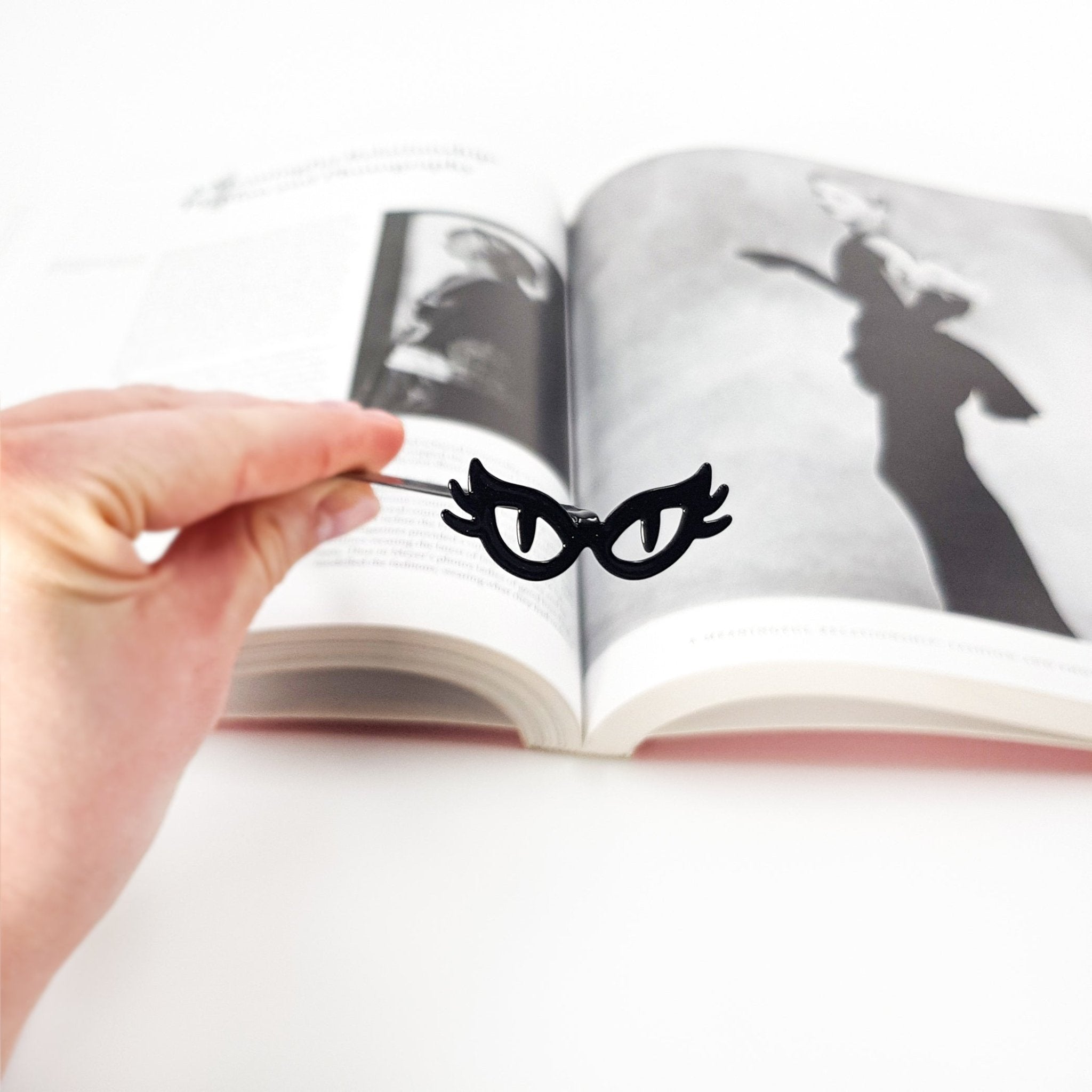 A hand holds a slim black metal bookmark shaped like cat’s eye glasses above an open book, with softly blurred pages in the background, highlighting the playful masquerade-style design against a clean white setting.