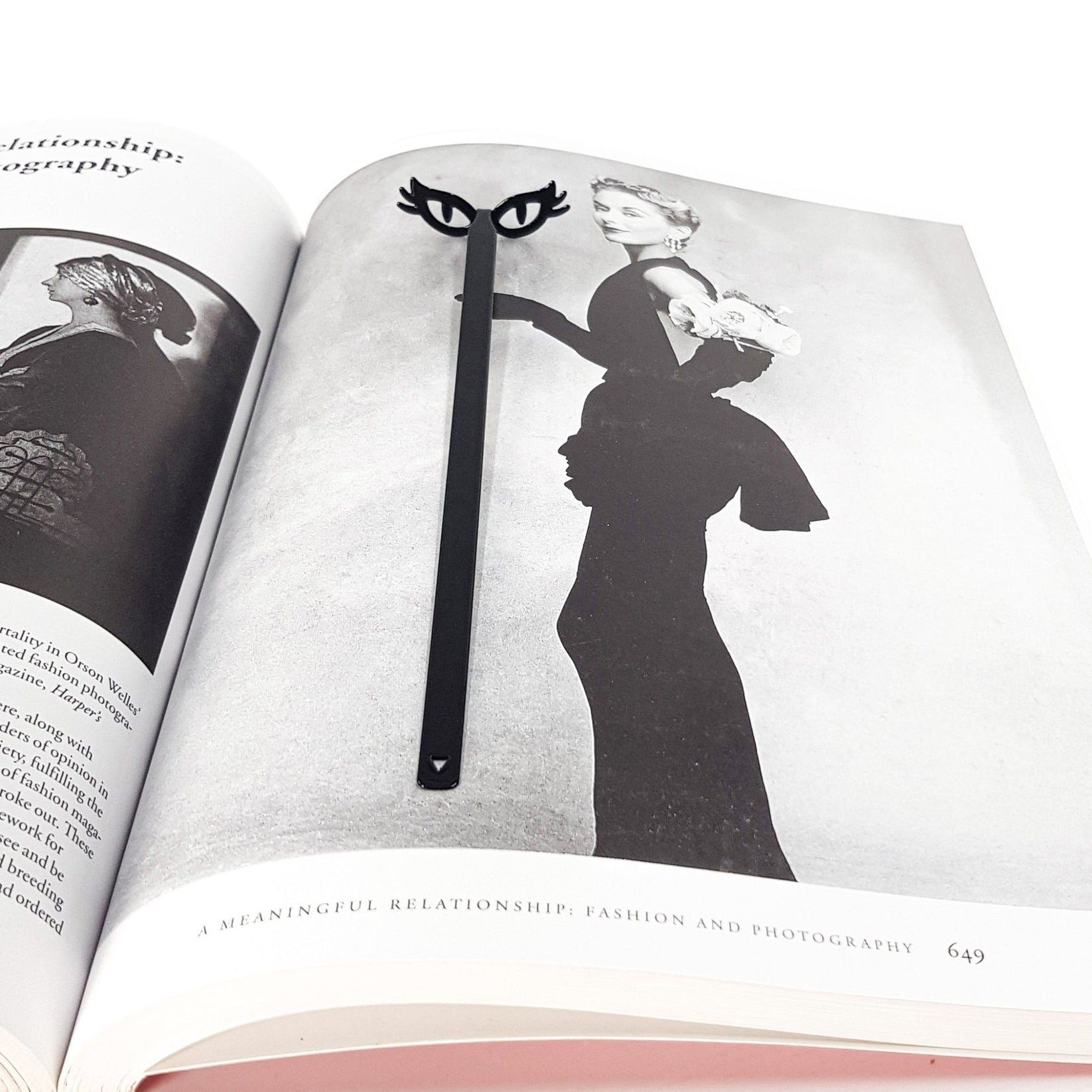Black metal cat’s eye glasses bookmark resting inside an open art book, aligned with a vintage fashion photograph, creating a playful masquerade mask effect on the page.