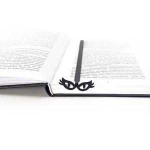 Black metal masquerade bookmark with cat’s eye glasses resting at the spine of an open book, minimalist bookish reading accessory