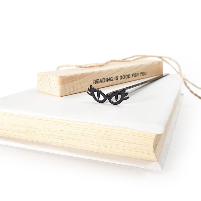 Black metal bookmark shaped like cat’s eye glasses resting on a closed book, with a wooden block reading “Reading is good for you” in the background.