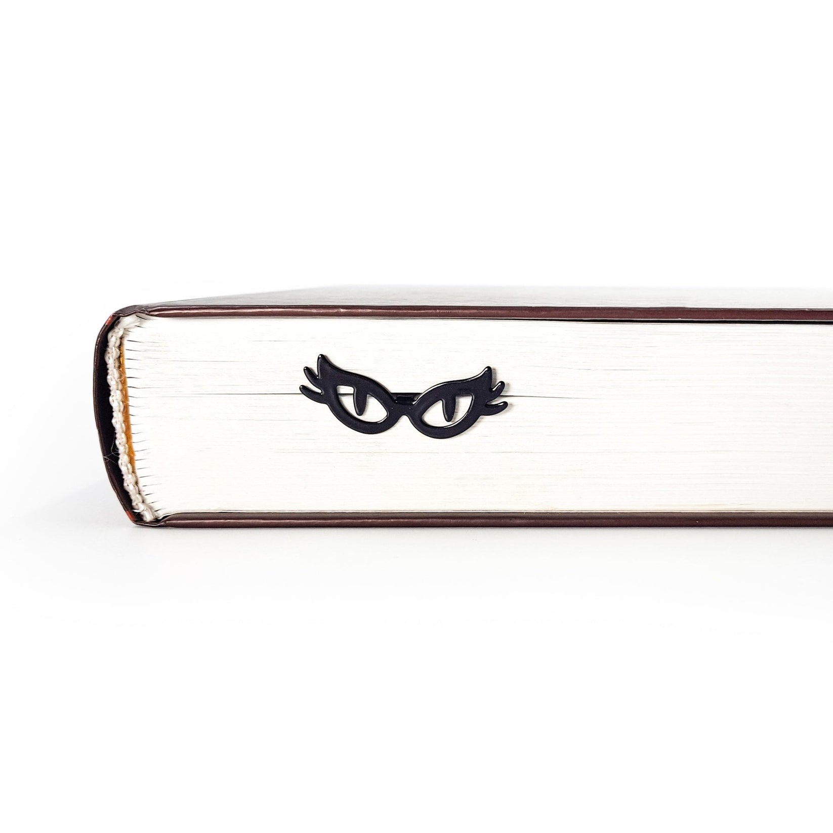 Black metal cat-eye glasses bookmark resting against the page edges of a closed book, creating a subtle peeking eyes effect for book lovers.