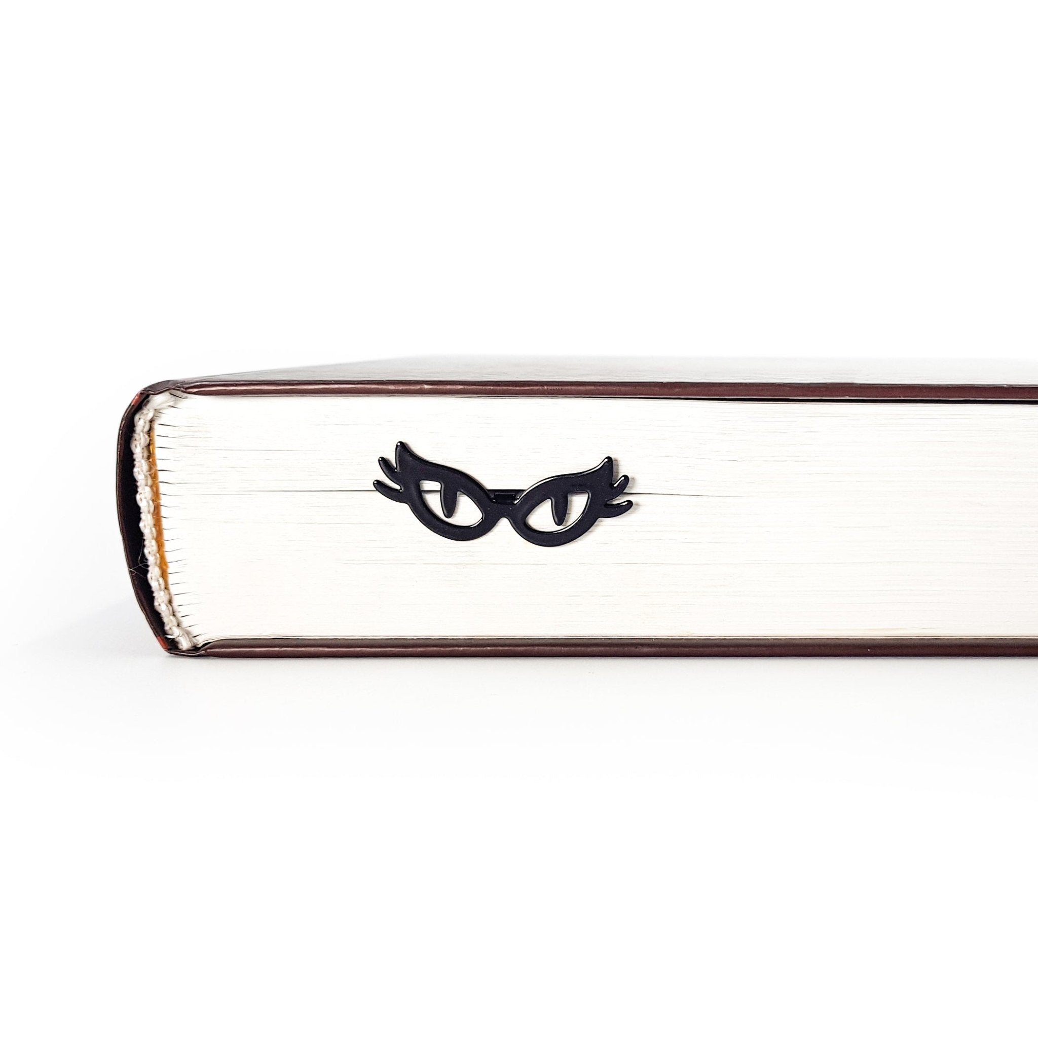 Black metal cat-eye glasses bookmark resting against the page edges of a closed book, creating a subtle peeking eyes effect for book lovers.