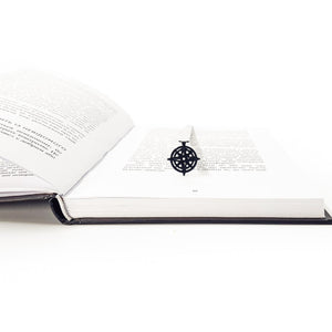 Compass rose metal bookmark resting across open book at angle, pendant centered on page