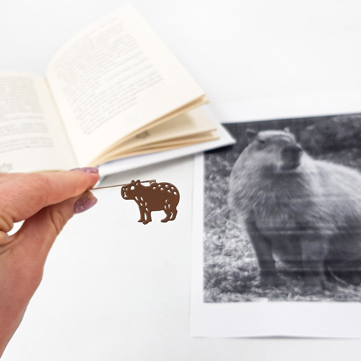 Capybara metal bookmark marking a page in an illustrated book, view 4