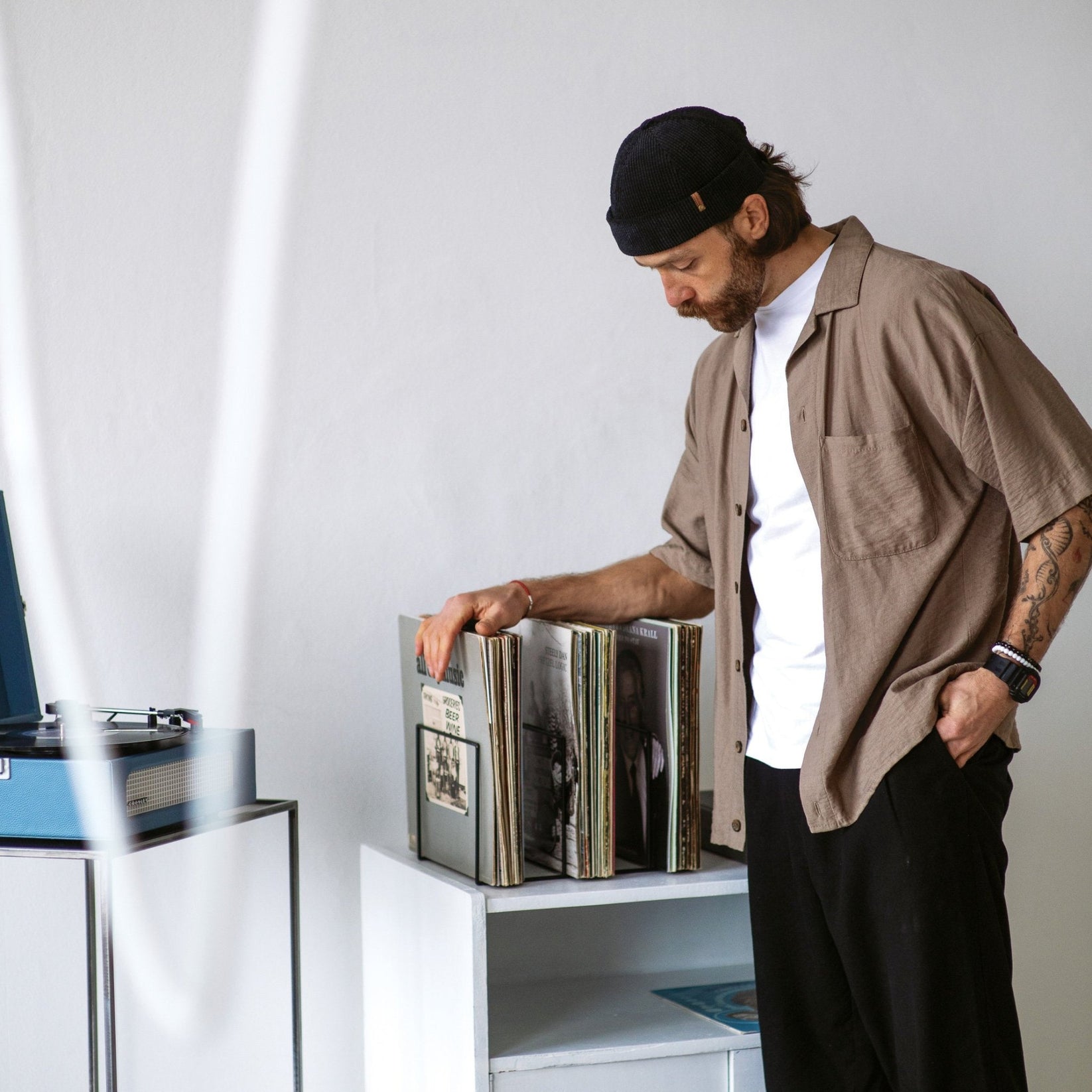 Man browsing vinyl records stored in a minimalist wire frame vinyl record stand, with a blue turntable visible on the left side of the image. Elegant Desktop Wire Frame Stand for Vinyl Records - Perfect for Organizing Your Collection - Atelier Article