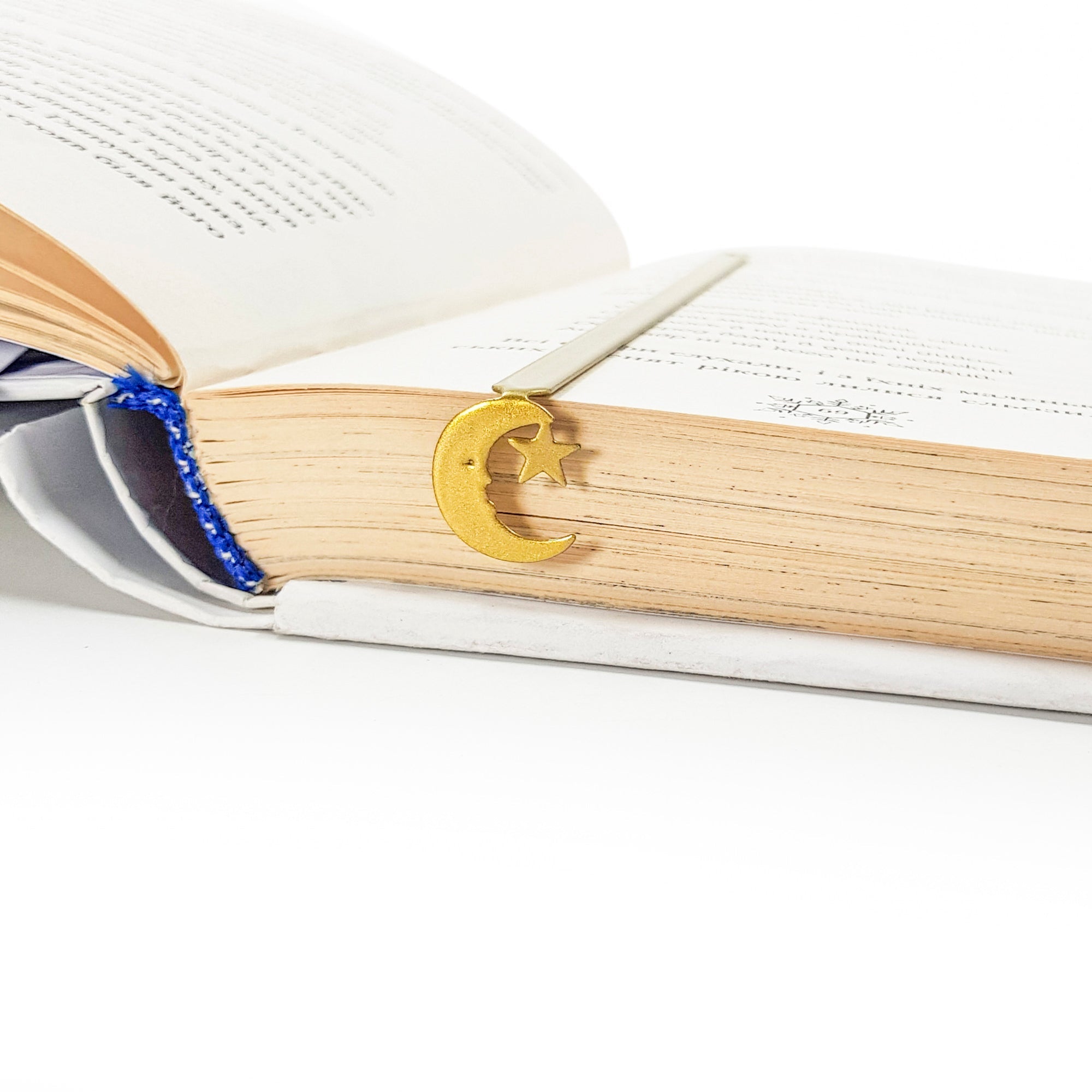 Gold crescent moon and star bookmark clipped to the pages of an open book with aged paper and visible blue thread binding
