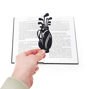 Hand holding a black metal golf bag bookmark above an open book, showing the laser-cut golf clubs silhouette and matte powder-coated finish used to mark a reading page.