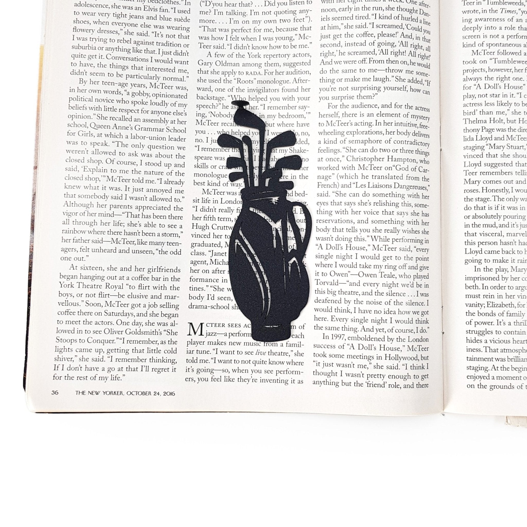 Black metal golf bag bookmark placed vertically between the pages of an open magazine, showing the slim profile and golf club silhouette against printed text.