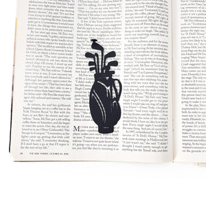 Black metal golf bag bookmark placed vertically between the pages of an open magazine, showing the slim profile and golf club silhouette against printed text.