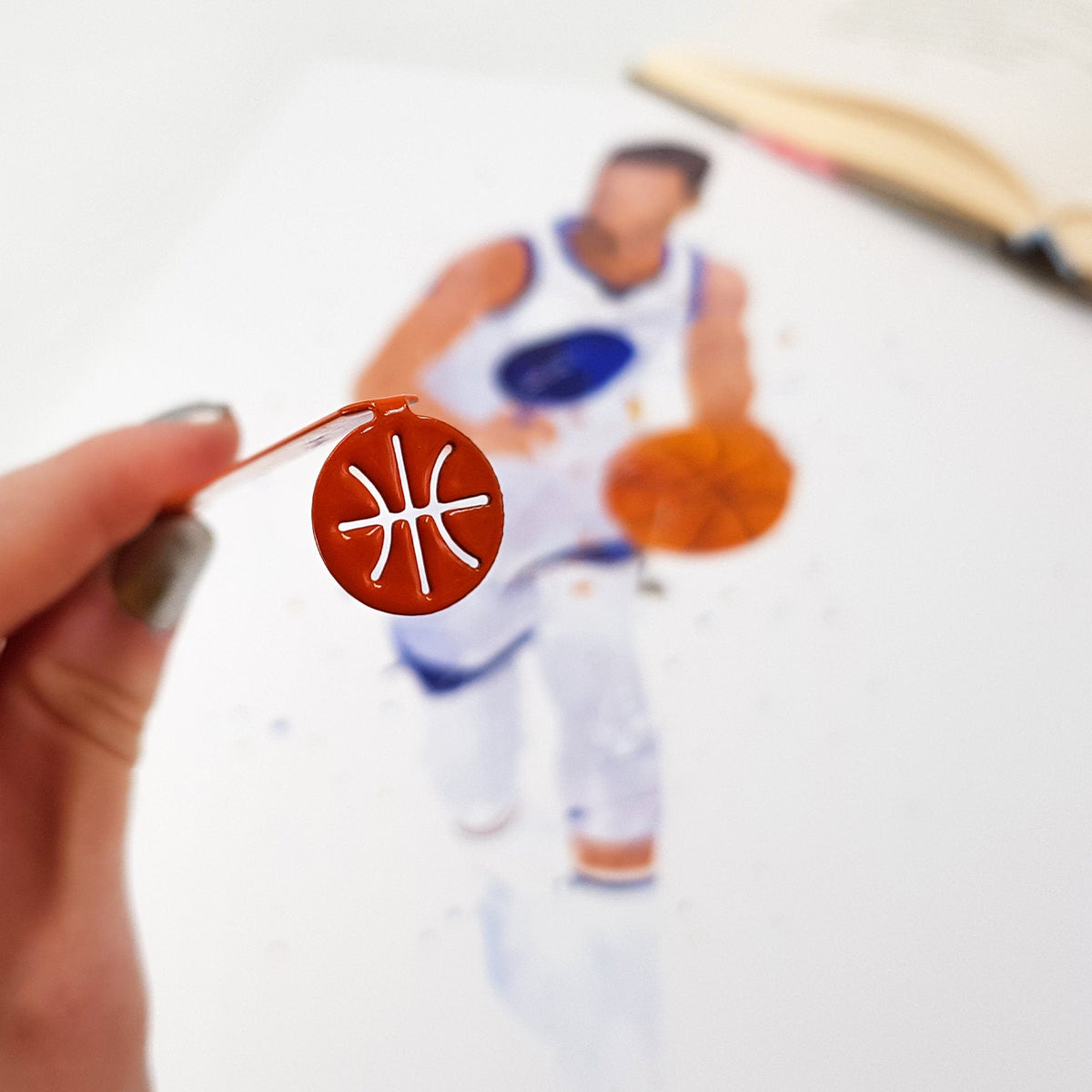Hoop Dreams Mark metal bookmark in red held in a hand above an open book