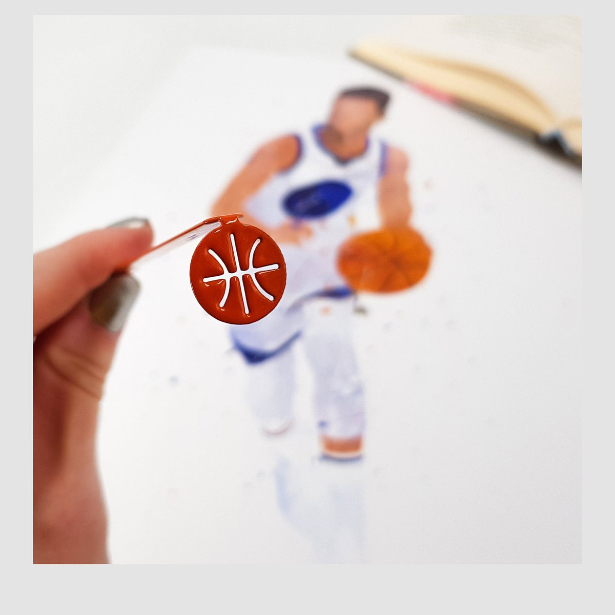 Hoop Dreams Mark metal bookmark in red held in a hand above an open book