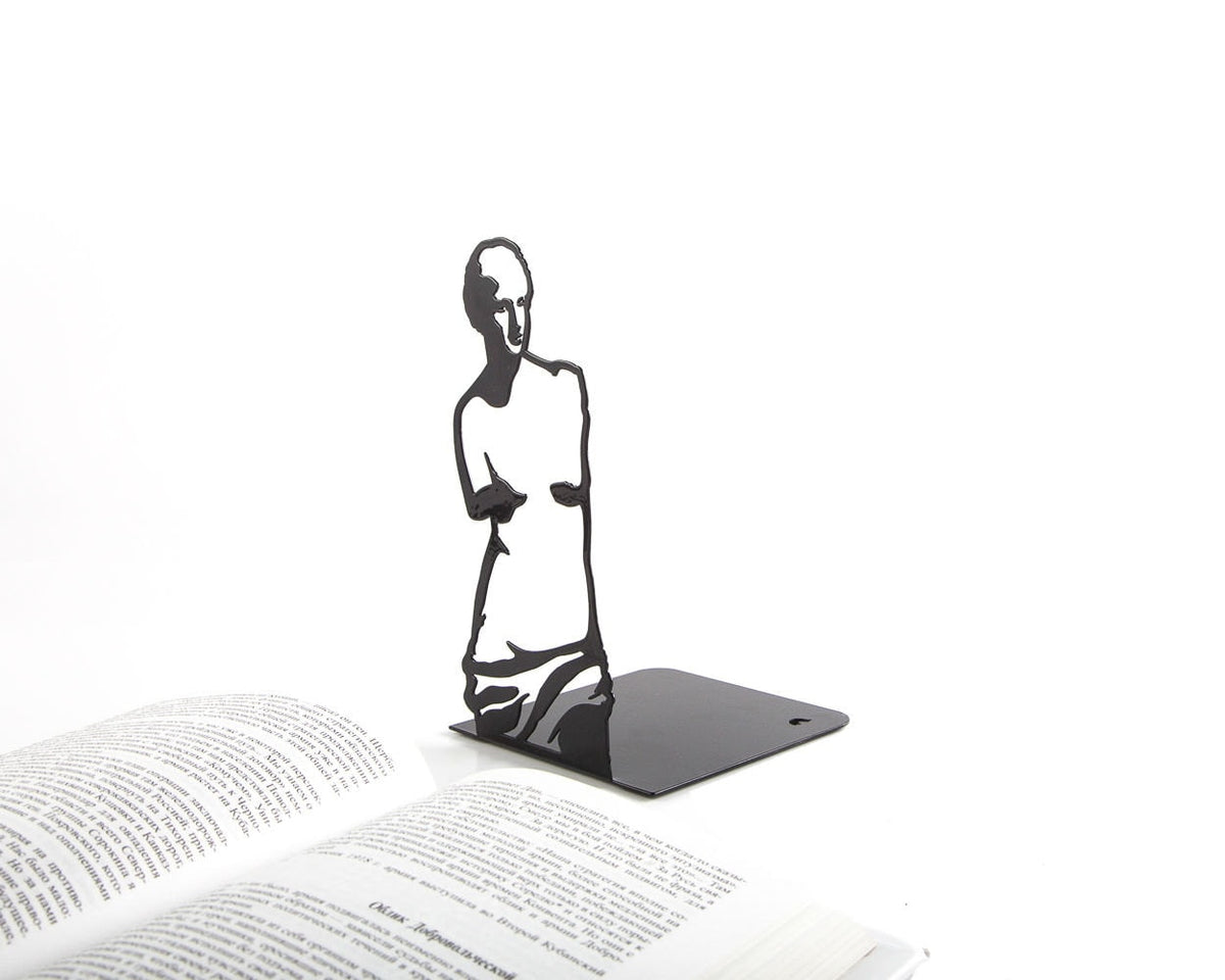 Black metal bookend in the shape of Venus de Milo silhouette, standing beside open books, elegant and artistic home library accessory.