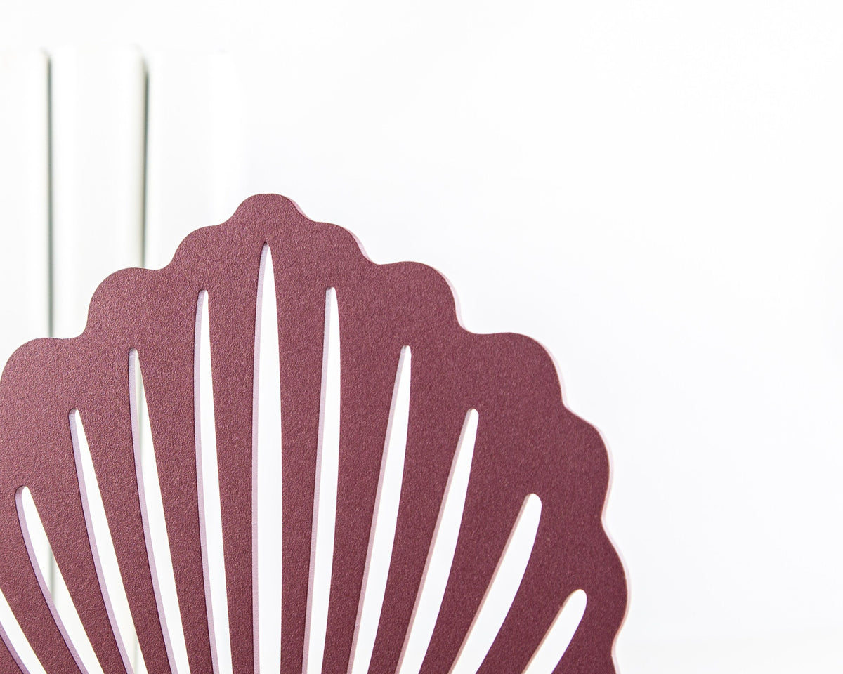 Close-up detail of the top part of a red metal seashell-shaped bookend with a matte finish, shown against a white background with books behind it.