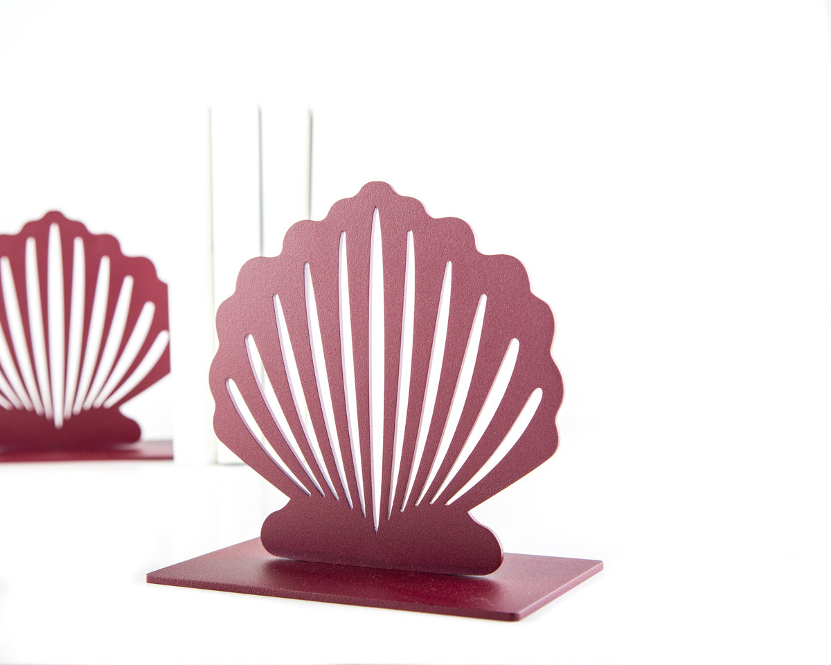 Close-up of a red metal seashell-shaped bookend with a modern cutout design, standing on a white surface with books and another matching bookend in the background.