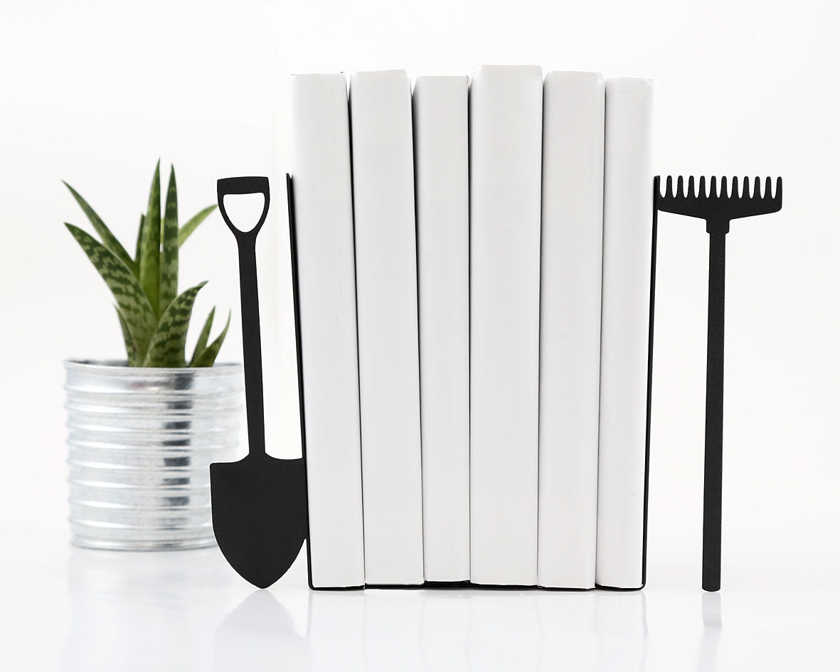 Pair of black metal bookends shaped like a garden shovel and rake, holding white books upright – perfect gift for gardeners, nature lovers, or outdoor enthusiasts. Minimalist design adds rustic charm and functional style to shelves or desks. Great for home libraries, garden rooms, or eco-inspired decor themes. Designed and made by Atelier Article on Etsy. Not mass-produced.