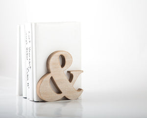 Natural wood ampersand symbol standing on white surface with stacked books in background, minimalist decorative accent for modern shelf or desk.