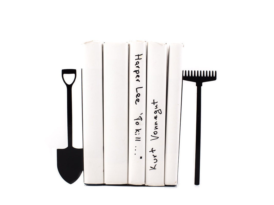 Metal bookends shaped like a shovel and a rake, holding white-covered books upright – minimalist garden tool design perfect for gardeners, nature enthusiasts, or rustic-themed home decor. Durable and playful shelf accessory for book lovers.