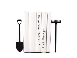 Metal bookends shaped like a shovel and a rake, holding white-covered books upright – minimalist garden tool design perfect for gardeners, nature enthusiasts, or rustic-themed home decor. Durable and playful shelf accessory for book lovers.