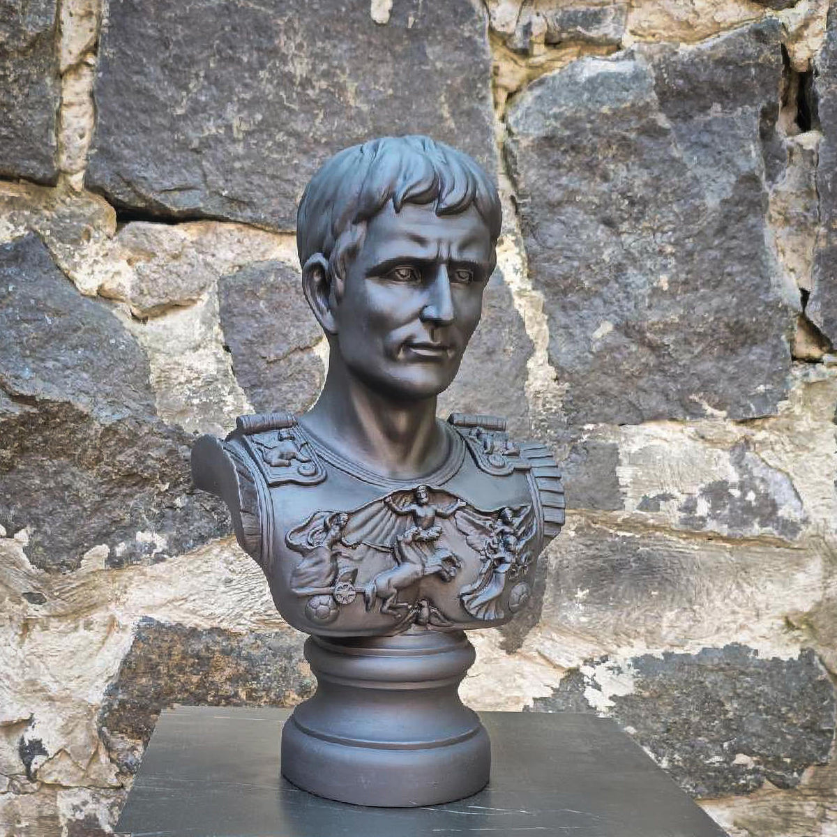 A dark gray plaster bust sculpture of a Roman gEmperor Julius Caesar in military armor and a lion emblem, displayed on a pedestal in front of a rustic stone wall.