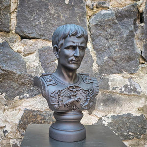 A dark gray plaster bust sculpture of a Roman gEmperor Julius Caesar in military armor and a lion emblem, displayed on a pedestal in front of a rustic stone wall.