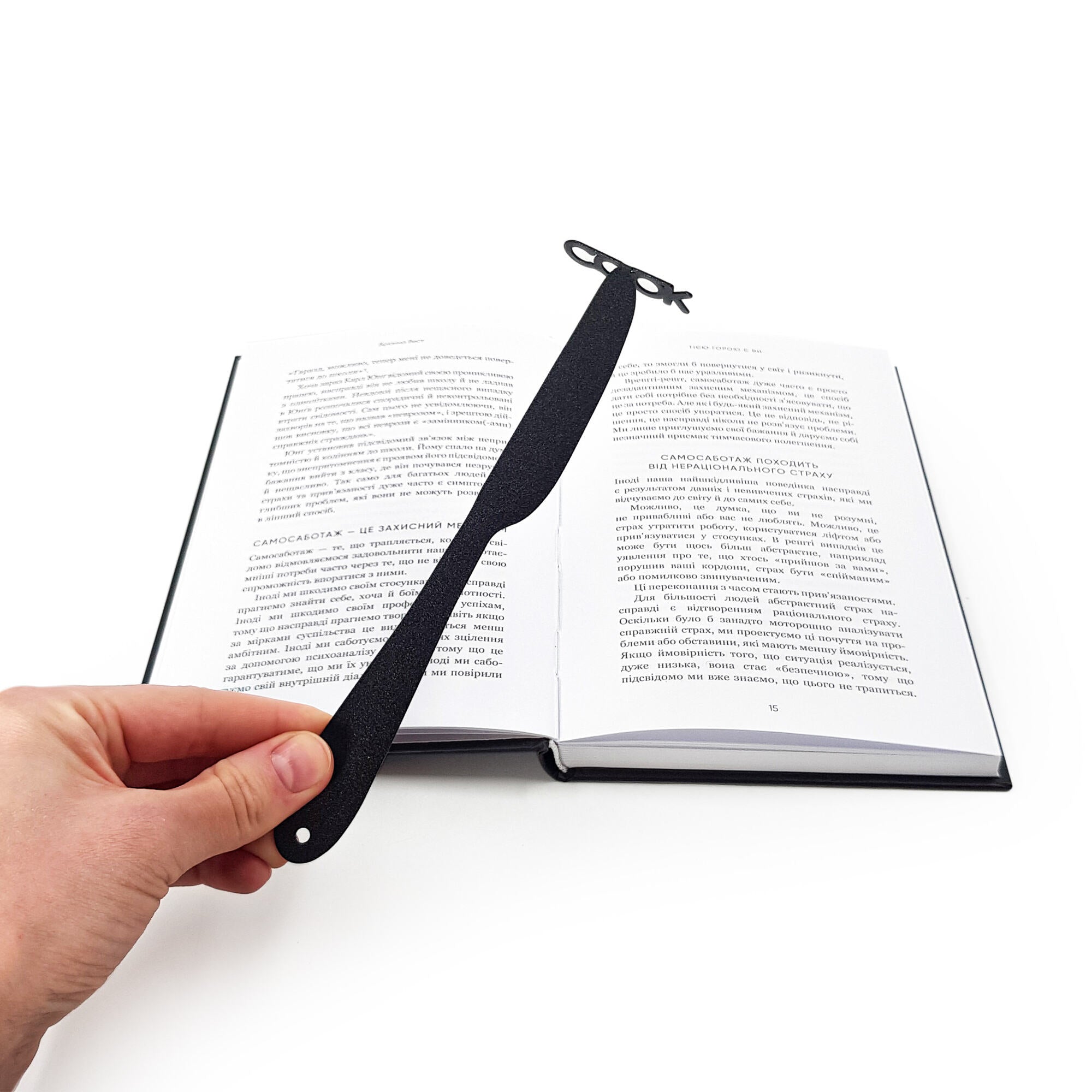 Hand holding a black knife-shaped COOK metal bookmark above an open book with Ukrainian text, pages spread flat on a white surface.