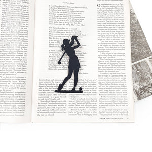 Black metal lady golf player bookmark placed on an open magazine page, showing the slim silhouette marking the text without covering it