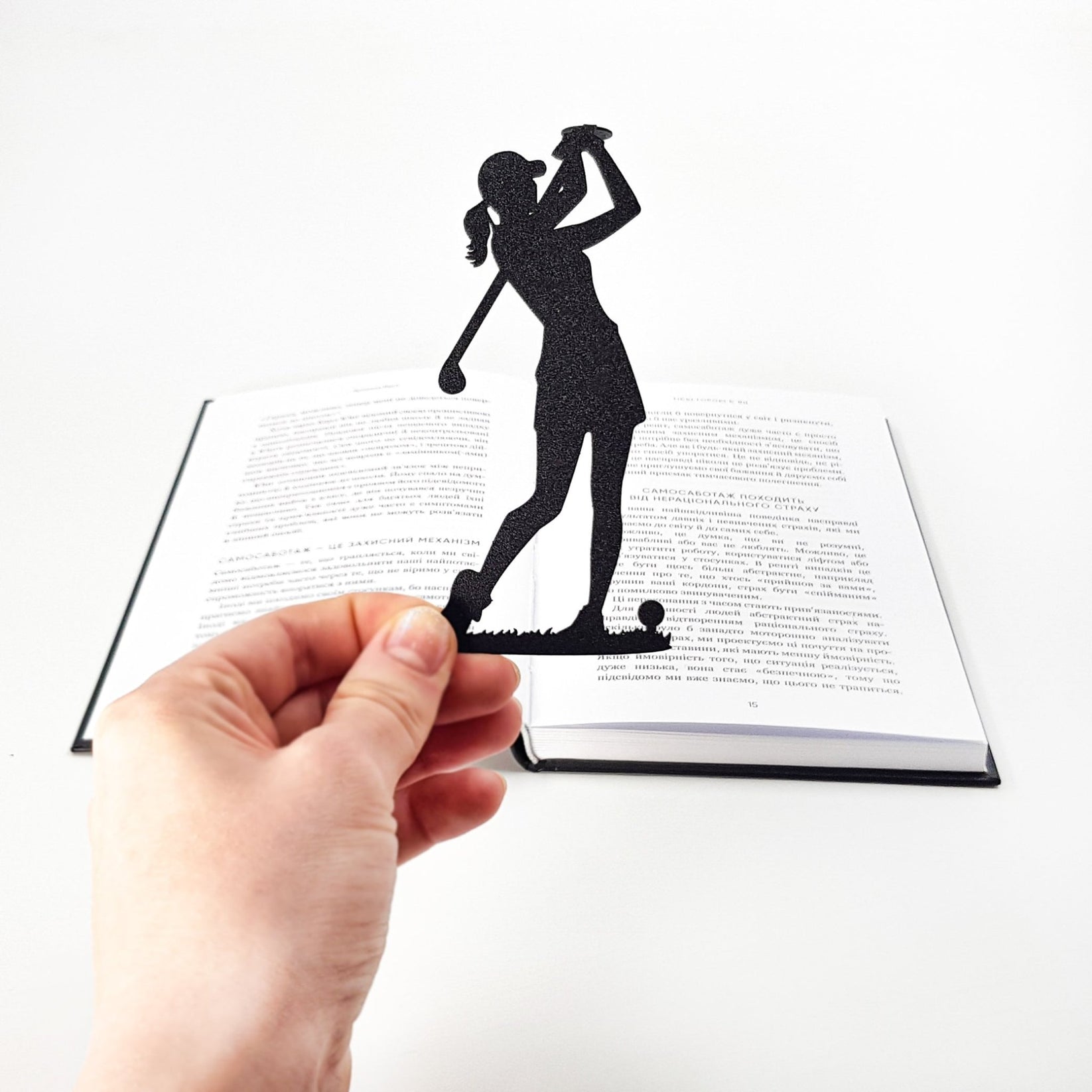 Hand holding a black metal lady golf player bookmark above an open book, demonstrating how the slim bookmark fits inside the pages