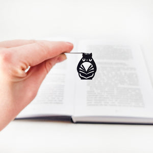 Hand holding a black laser cut owl metal bookmark in front of an open book, highlighting the slim design and detailed owl silhouette.