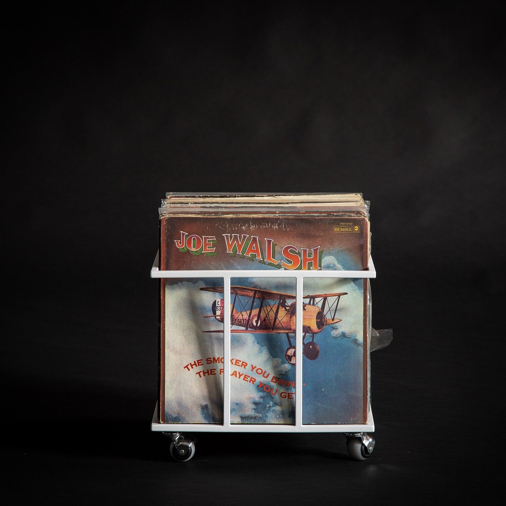 White metal record storage crate on wheels, holding vintage vinyls with a Joe Walsh album in front, displayed against a black background.
