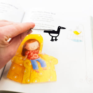 Hand holding a minimalist black metal bird bookmark above an illustrated book page, showing the bird silhouette and slim metal spine.