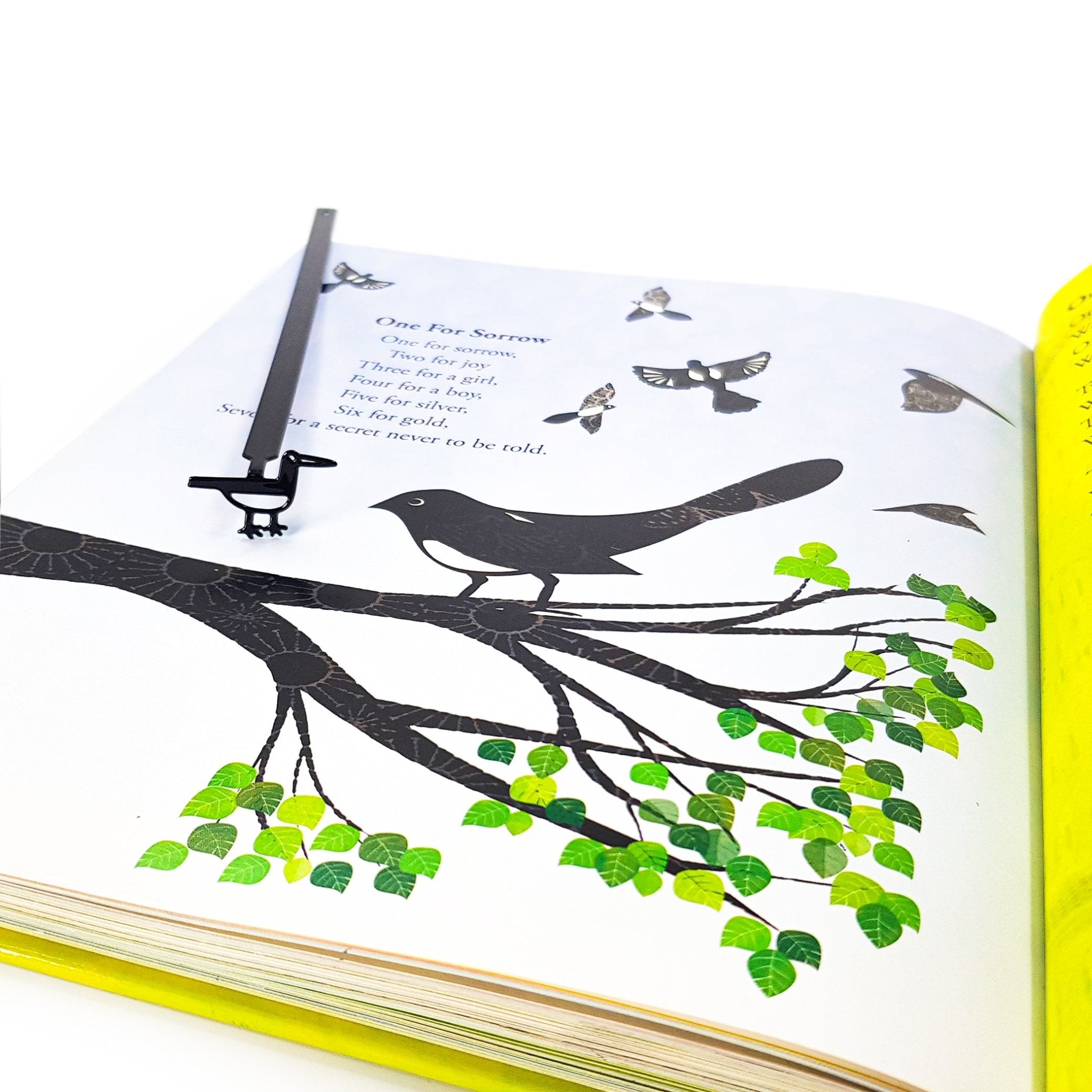 Minimalist black metal bird bookmark marking a page in an illustrated book with bird artwork, showing how the bookmark aligns with the page design.