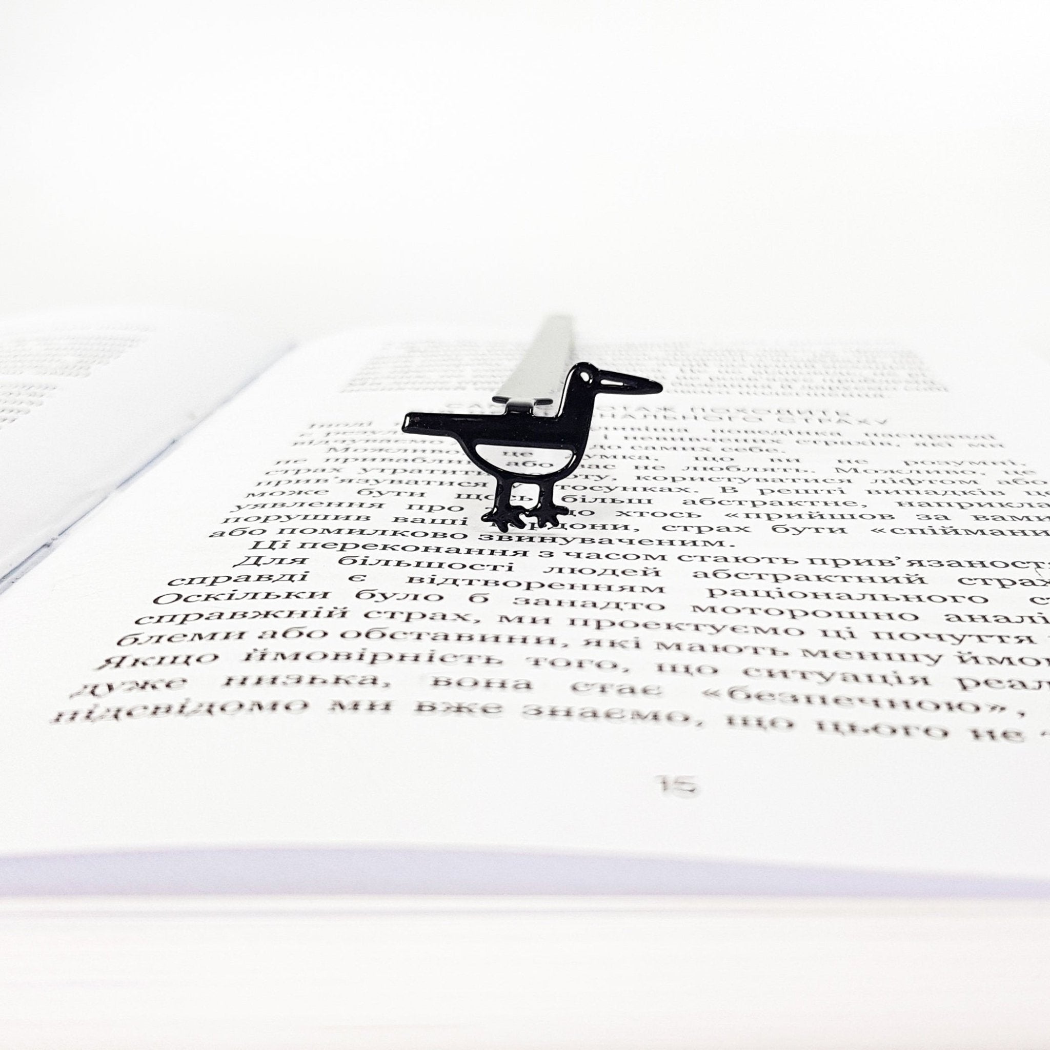 Minimalist black metal bird bookmark standing on an open book page, shown from the front to highlight the bird shape and slim metal spine.