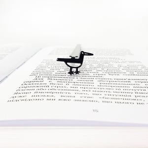 Minimalist black metal bird bookmark standing on an open book page, shown from the front to highlight the bird shape and slim metal spine.