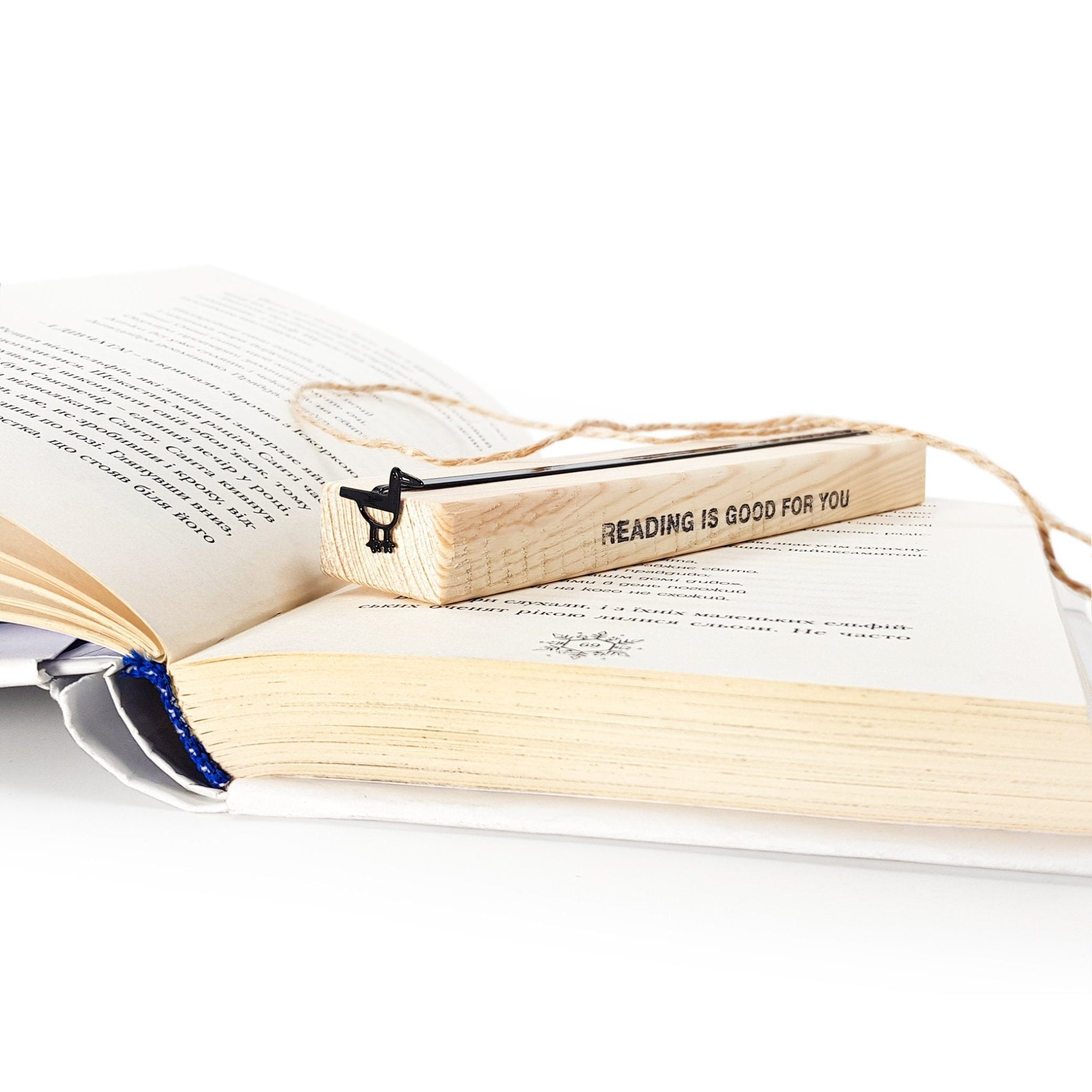 Minimalist black metal bird bookmark displayed on a wooden block engraved “READING IS GOOD FOR YOU,” placed on an open book to show gift presentation and scale.