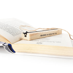 Minimalist black metal bird bookmark displayed on a wooden block engraved “READING IS GOOD FOR YOU,” placed on an open book to show gift presentation and scale.