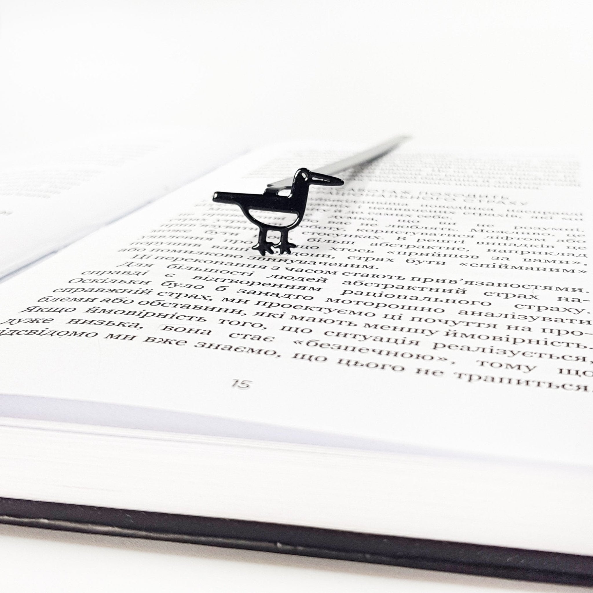 Minimalist black metal bird bookmark standing on an open book page, viewed from a low angle to show the slim profile and page-marking position.