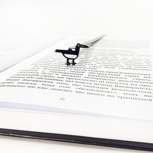 Minimalist black metal bird bookmark standing on an open book page, viewed from a low angle to show the slim profile and page-marking position.