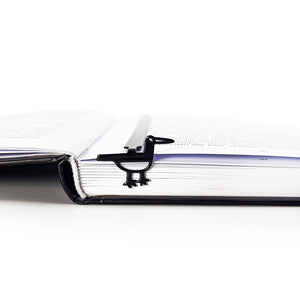 Minimalist black metal bird bookmark clipped onto the pages of an open book, shown from the side to illustrate how it holds place securely.