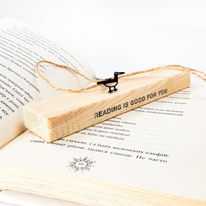 Minimalist black metal bird bookmark resting on a wooden block engraved “READING IS GOOD FOR YOU,” placed on an open book.