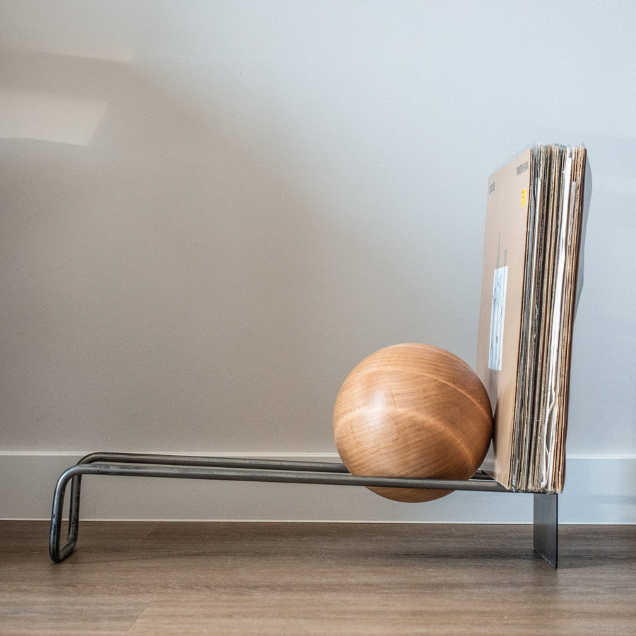 Black metal record and book stand with a wooden ball holding records beside a pale wall