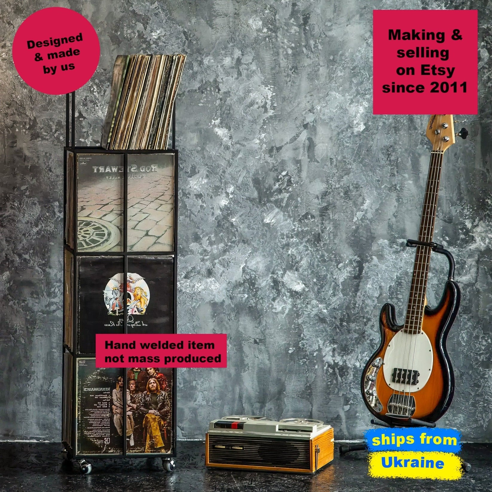 Tall black vinyl record holder on wheels, with four compartments, displayed against a textured gray wall beside a vintage bass guitar and retro tape recorder.