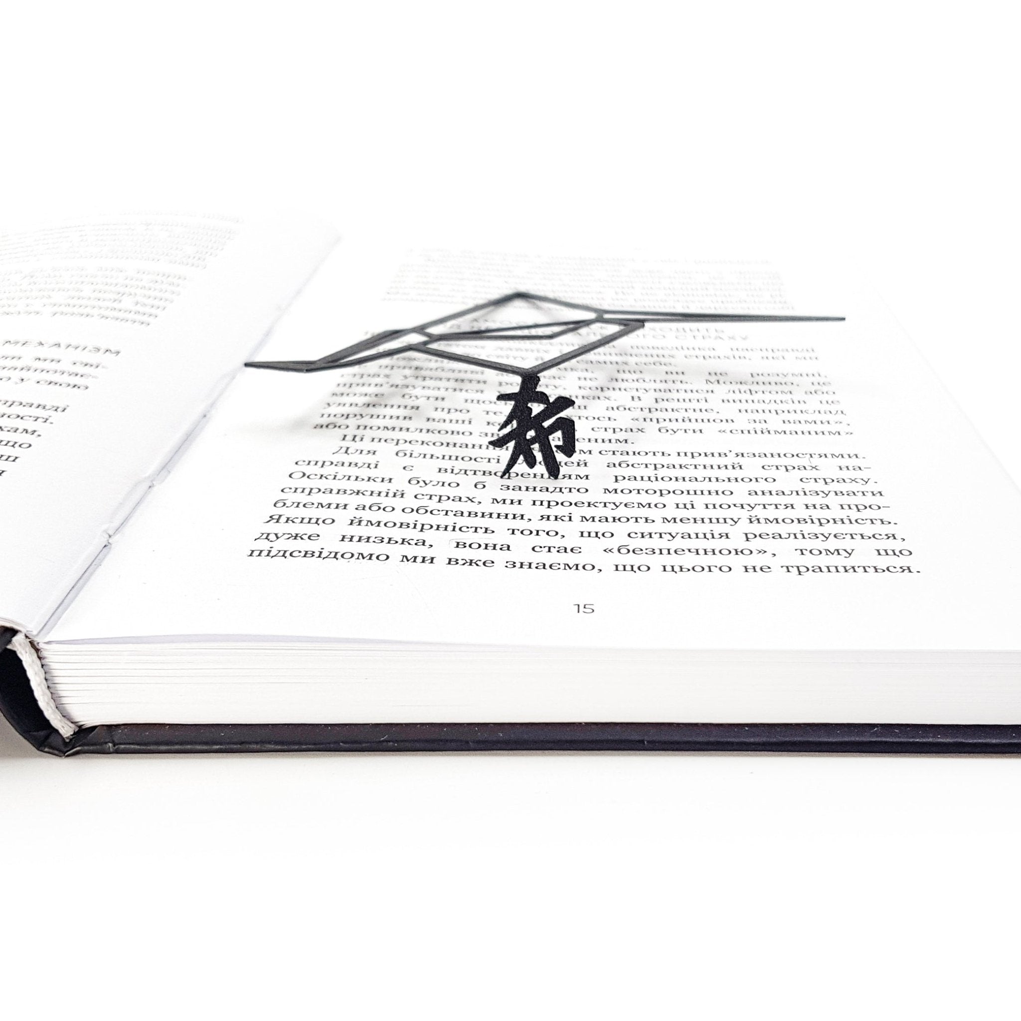 Low-angle close-up of a black metal origami crane bookmark lying flat across the pages of an open book, with its thin geometric form hovering slightly above the printed text.