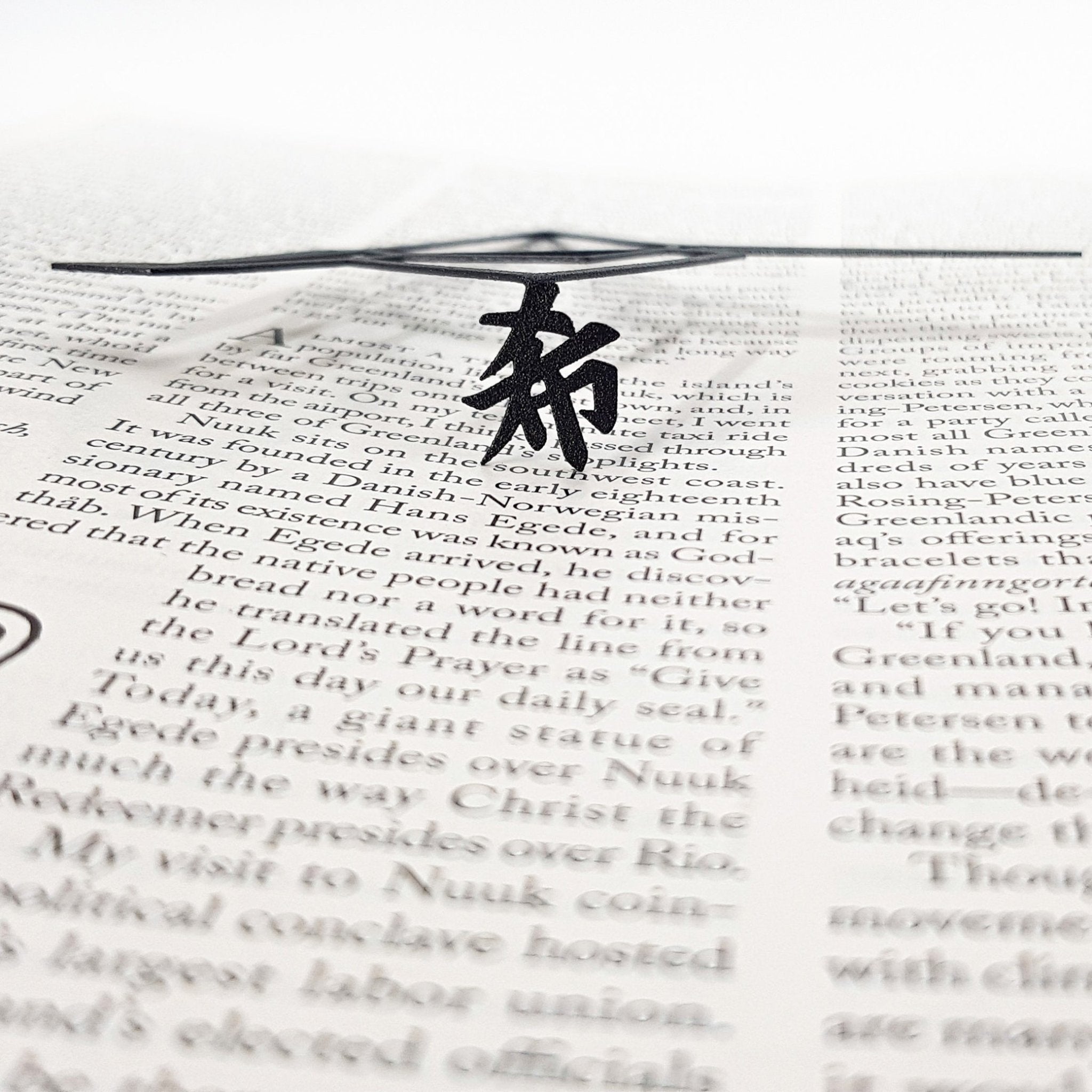 Black metal bookmark resting across an open book, showing a Zen kanji symbol beneath a thin geometric metal design.