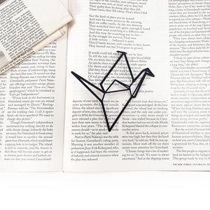 Black metal origami bird bookmark resting across an open book, highlighting a minimalist geometric silhouette against printed text.