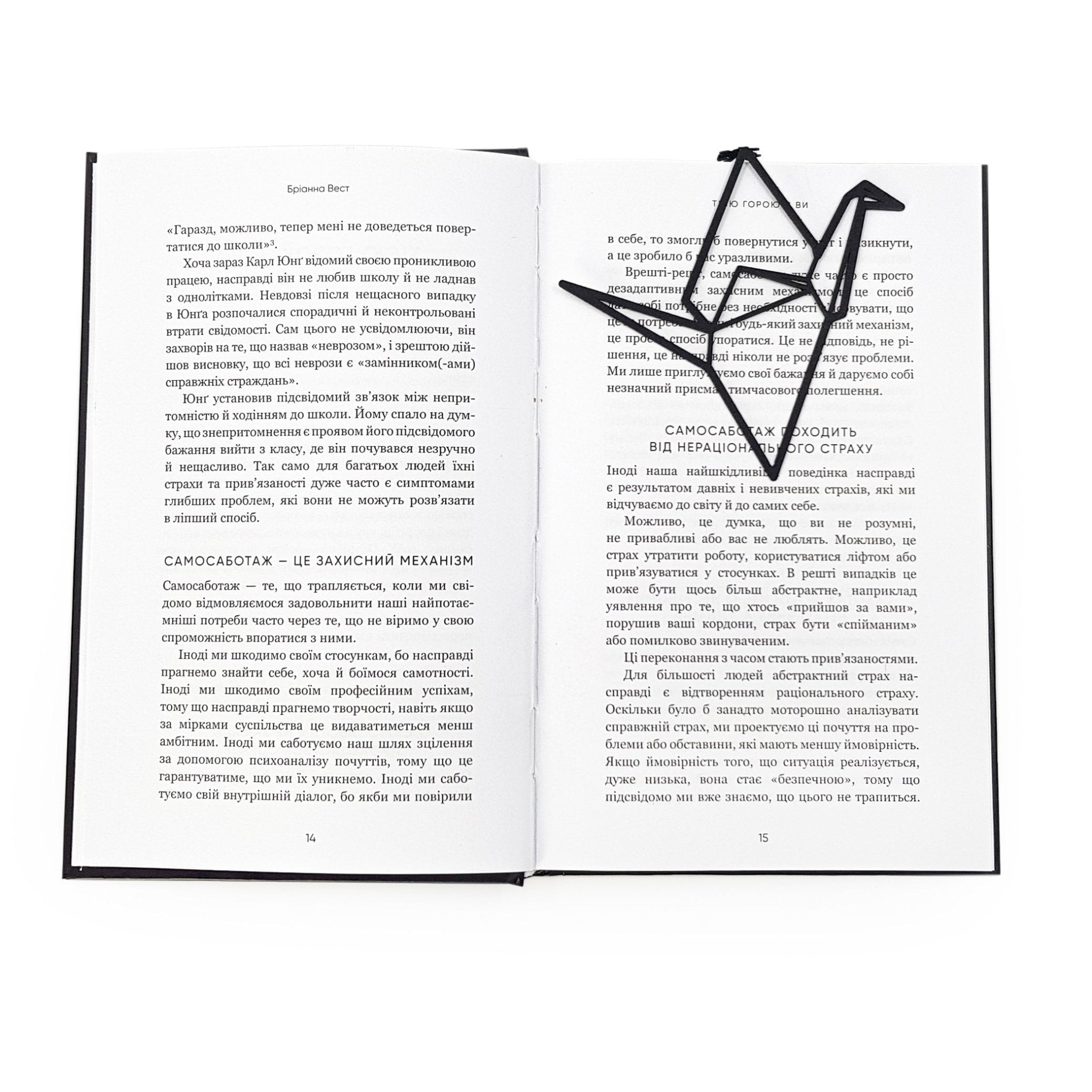 Black metal origami crane bookmark resting diagonally on the pages of an open book, highlighting its minimalist geometric design against printed text.