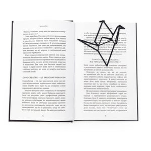 Black metal origami crane bookmark resting diagonally on the pages of an open book, highlighting its minimalist geometric design against printed text.