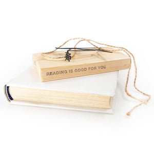 Black metal bookmark with a Zen kanji symbol resting on a wooden block engraved “Reading is good for you,” placed on a closed book.