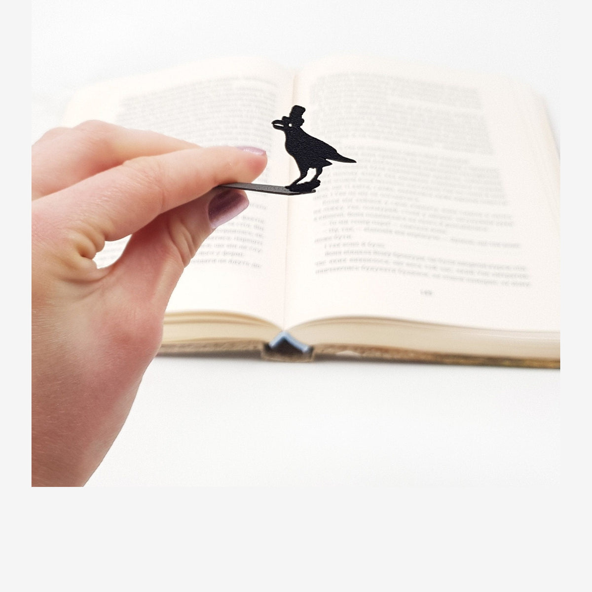 Raven in High Hat metal bookmark held in a hand above an open book